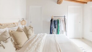 Ein Schlafzimmer mit Holzbalkendecke, einem Bett mit beigefarbener Bettwäsche, einem Kleiderschrank und einem Kleiderständer mit verschiedenen Kleidungsstücken.