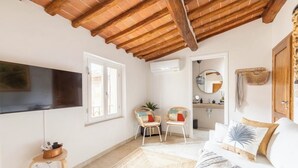 Ein Schlafzimmer mit Holzboden, einem an der Wand montierten Flachbildfernseher, zwei Korbstühlen, einem Bett mit weißer Bettwäsche und einem kleinen Tisch mit einem Korb.