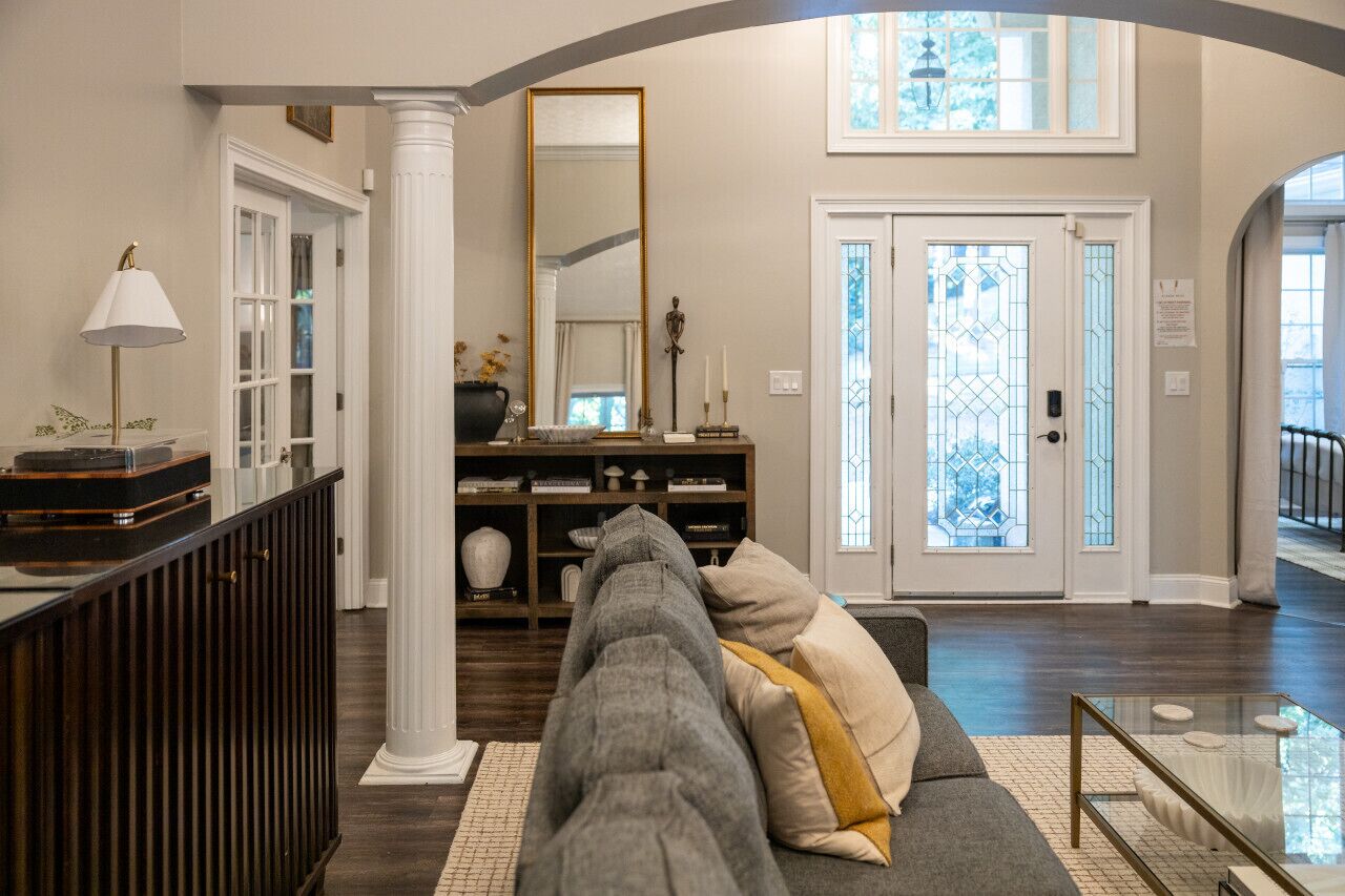 A living room with a dark wood floor, a grey sofa with a mustard throw pillow, a glass coffee table, and a bookshelf.