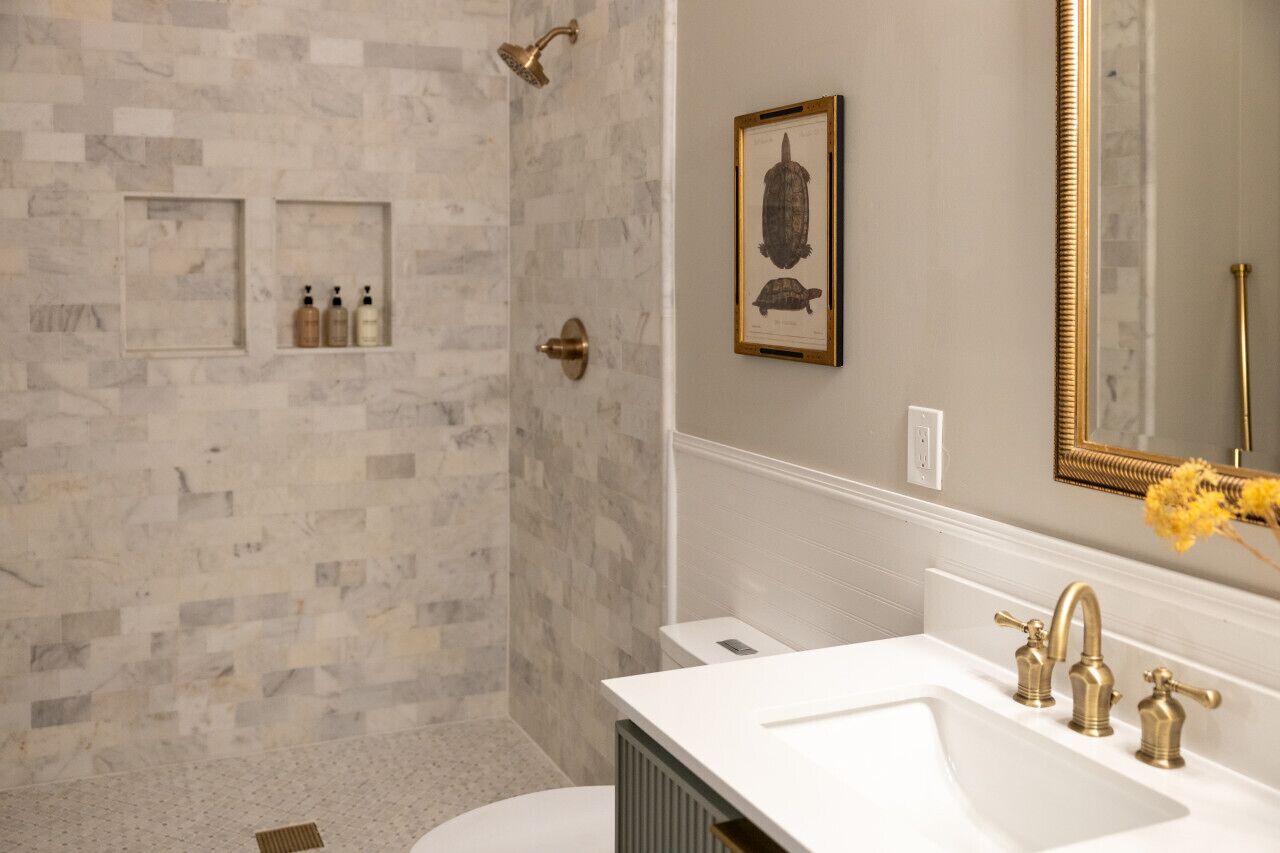 A bathroom with a marble shower wall, a white sink with gold faucets, and a framed turtle picture on the wall.