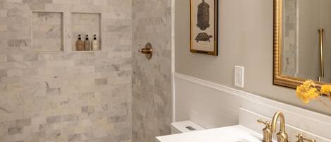 A bathroom with a marble shower wall, a white sink with gold faucets, and a framed turtle picture on the wall.