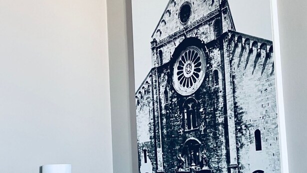 A hotel room with a large black and white photo of a historic building on the wall, a bedside table with a lamp, and a neatly made bed. - Full house (Bari)