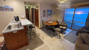 A kitchen with a sink, a coffee maker, and a dining area with a table and chairs. There are framed pictures on the wall and a view of the ocean through a large window.