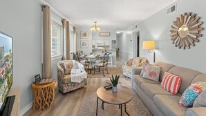 Een woonkamer met een bank, fauteuils, een salontafel en een televisie. Er is een eethoek met een tafel en stoelen, en een keuken op de achtergrond.