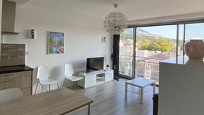 A modern living room with a TV, white chairs, a wooden table, and a large window with a city view. - Modern studio with breathtaking view close to the train station (Aix-les-Bains)