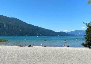 A pebbled beach with people sunbathing and swimming, surrounded by mountains and clear blue water.
