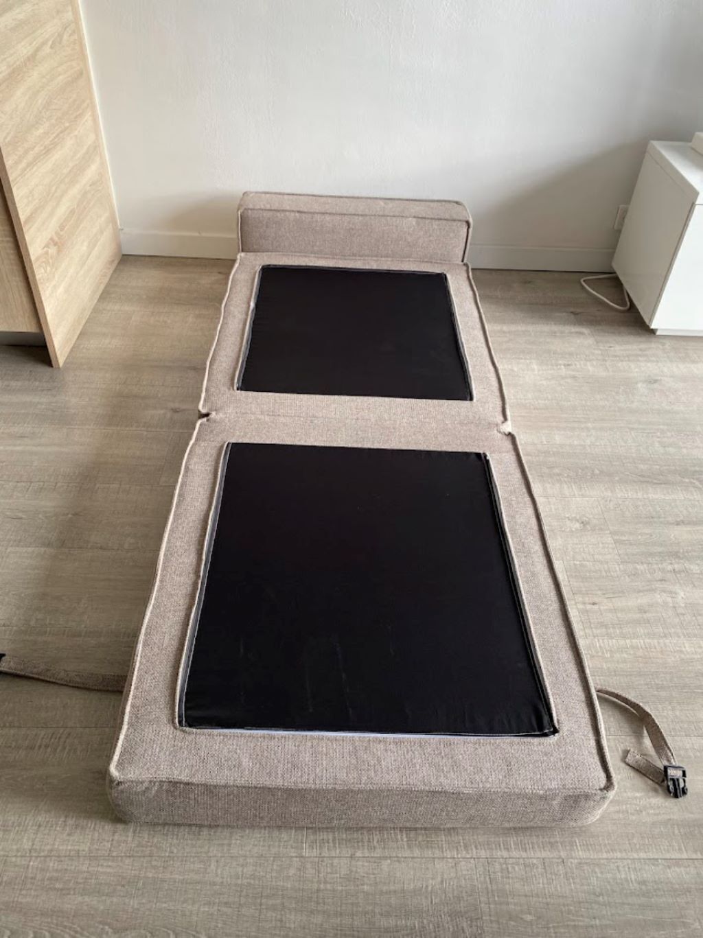 Ein ausklappbares Sofa mit grauem Stoffbezug und schwarzen Kissen, stehend auf einem Holzboden vor einer weißen Wand.