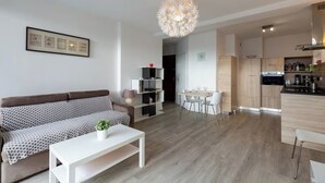 A modern living room with a sofa, coffee table, and dining area with chairs. There is a kitchen with a stove, oven, and cabinets. A chandelier hangs from the ceiling. - Modern studio with breathtaking view close to the train station (Aix-les-Bains)