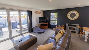 A living room with a sofa, ottoman, television, fireplace, and a large clock on the wall. - Remarkable  Rhoboro Retreat (Twizel)