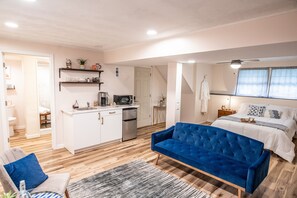 Un espacio habitable compacto con kitchenette, un sofá azul, una cama con ropa de cama blanca y un ventilador de techo.