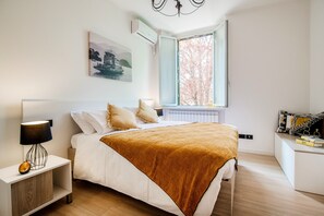 A modern bedroom with a large bed, a bedside table, a nightstand, a window with a view of trees, and a painting on the wall. - MyHomeInComo - Promenade (Cernobbio)