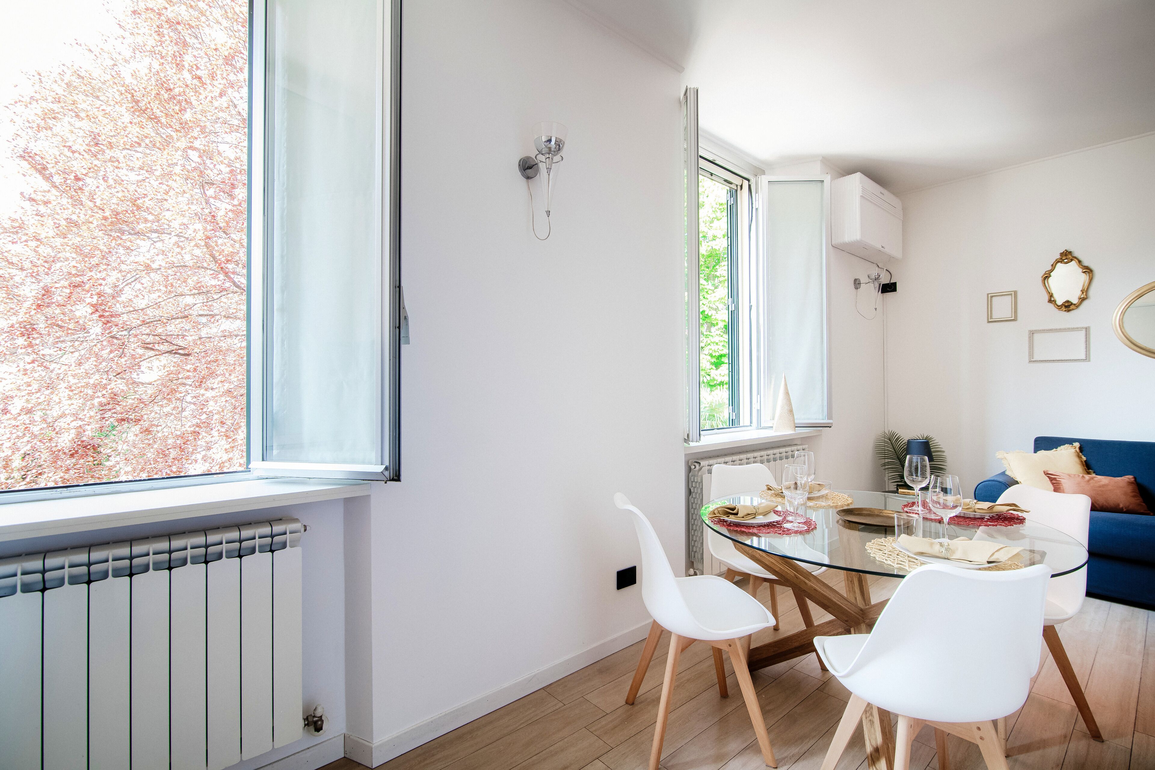 Un espace de salle à manger moderne avec une table en verre, des chaises blanches et une vue sur les arbres à travers une grande fenêtre.