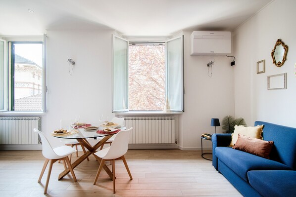 A modern living room with a dining area, a blue sofa, and a large window with a view of trees. - MyHomeInComo - Promenade (Cernobbio)