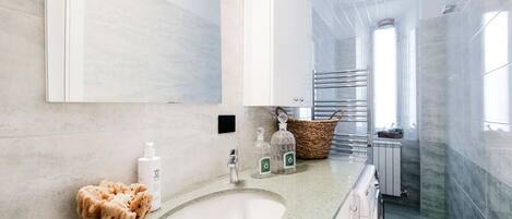 A modern bathroom with a large mirror, a green countertop, and a white sink. There are toiletries on the counter and a basket in the background.
