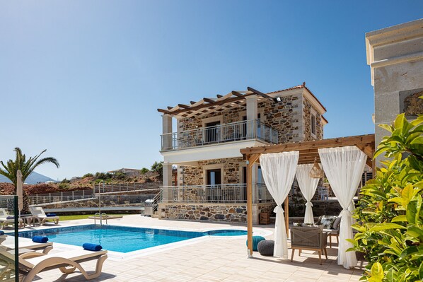 A poolside area with lounge chairs, a pergola with white curtains, and a stone building with a balcony. - Agapi Villa II, a Seaside Retreat with Pool & BBQ, Walking distance to beach (Rethymno)