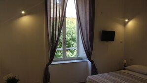 Een slaapkamer met een bed, een raam met gordijnen, een klein tafeltje met een plant en een aan de muur bevestigde televisie.