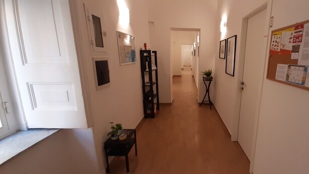 Een smalle gang met witte wanden, een houten vloer en een deur aan de rechterkant. Er hangen afbeeldingen in lijsten aan de muren en een prikbord met mededelingen.