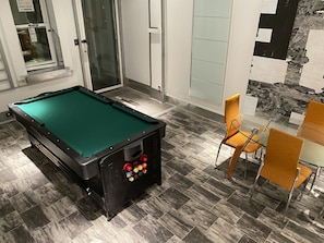 Una mesa de billar con superficie negra y fieltro verde, una puerta de cristal, una silla y una mesa con sillas.