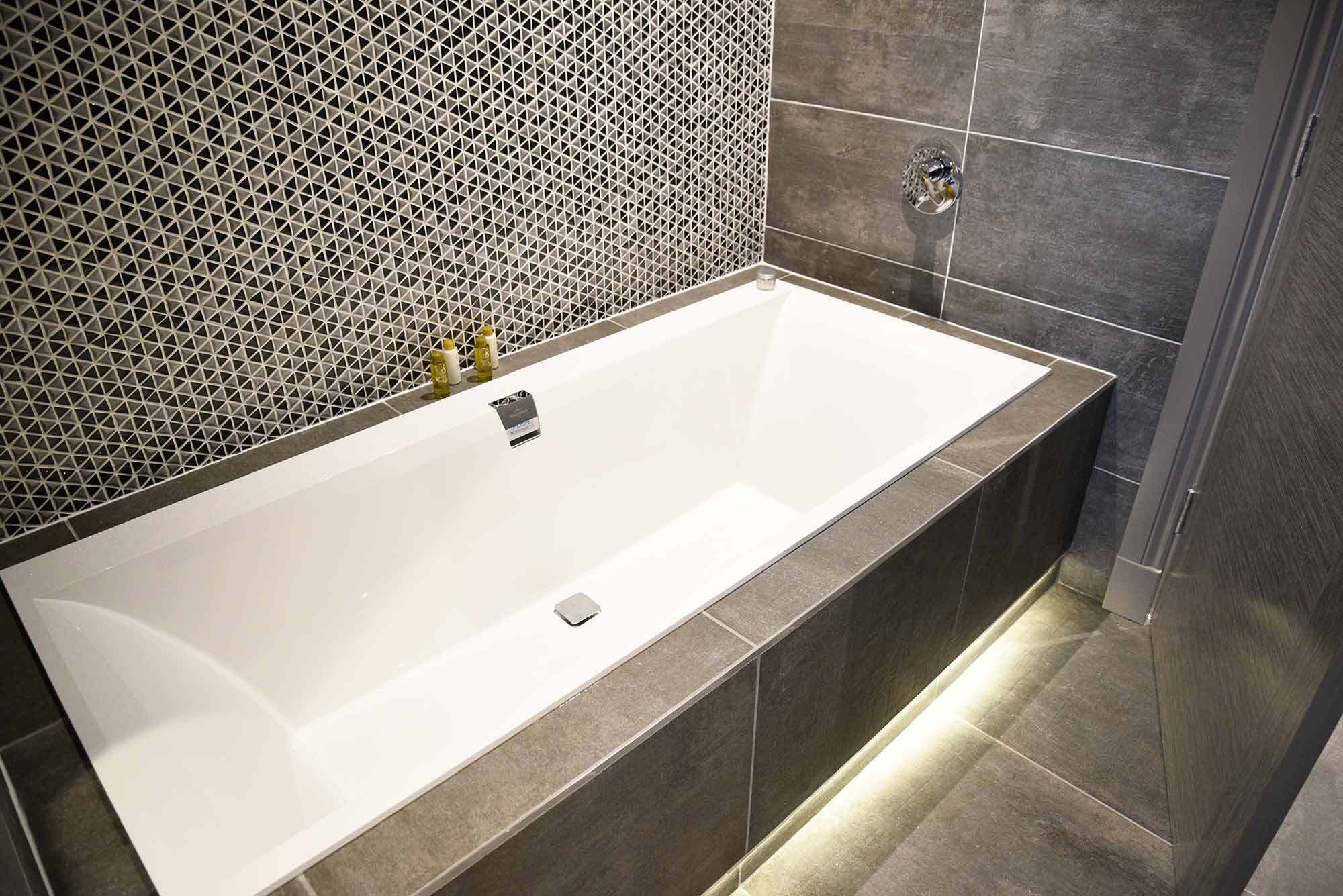 A modern bathroom with a rectangular bathtub, dark tiled walls, and a patterned tile backsplash.