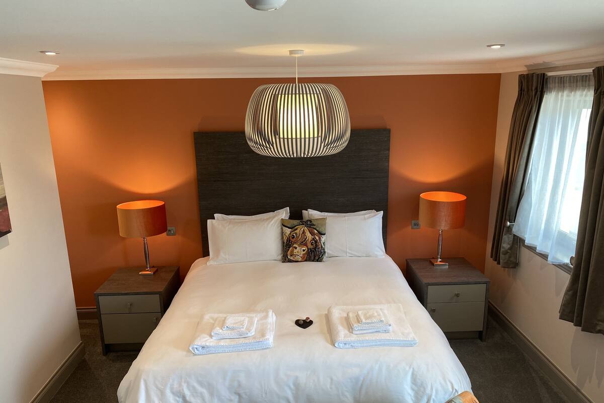 Ein Hotelzimmer mit einem großen Bett, zwei Nachttischlampen, einer modernen Chandelierleuchtre und einem Fenster mit Vorhängen.