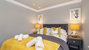 Un lit bien fait, avec une couverture jaune et des serviettes blanches, deux lampes de chevet et des œuvres d’art accrochées aux murs.