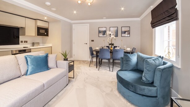 Un salon moderne comprenant un canapé, un fauteuil bleu, une table à manger avec des chaises et un coin cuisine équipé d’un four à micro-ondes et de placards.
