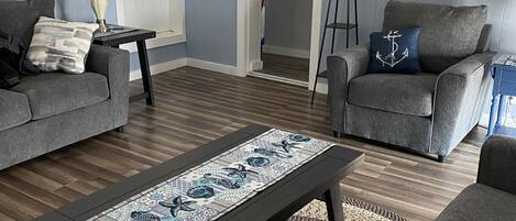A living room with two grey sofas, a coffee table with a patterned table runner, a floor lamp, and a woven rug.