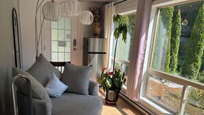 A modern living room with a grey sofa, a standing heater, a potted plant, and a view of greenery outside. - Entire Suite,Ocean,mountain view,jacuzzi in the Suite,next to Mount Maxwell park (Salt Spring Island)