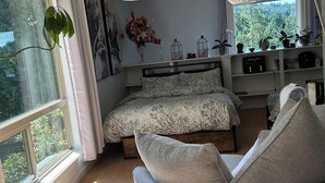 A bedroom with a bed, a chair, a window with curtains, a plant, and a shelf with various items. - Entire Suite,Ocean,mountain view,jacuzzi in the Suite,next to Mount Maxwell park (Salt Spring Island)