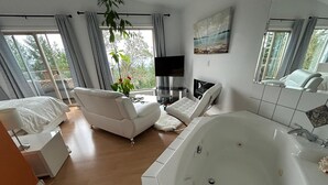 A modern bedroom with a large white bathtub, a sofa, a TV, and a view of the outdoors through large windows. - Entire Suite,Ocean,mountain view,jacuzzi in the Suite,next to Mount Maxwell park (Salt Spring Island)