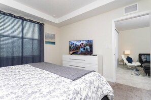 Un dormitorio con cama, televisor sobre un aparador, una silla y una puerta que da a otra habitación.