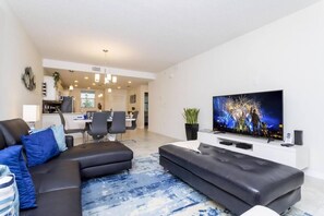 A modern living room with a black sectional sofa, a flat-screen TV, and a dining area in the background.
