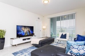 Ein modernes Wohnzimmer mit Flachbildfernseher, einer Couch, einem Couchtisch und einem Fenster mit Jalousien.