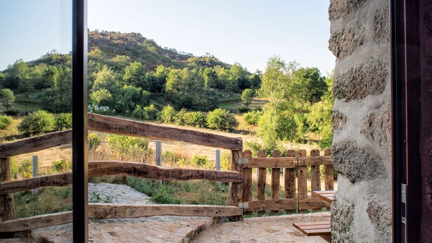 A stone pathway leading to a wooden bridge over a river, with a view of a forested hillside.
