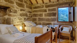 A stone-walled bedroom with two beds, a wicker chair, and a window with a mountain view.