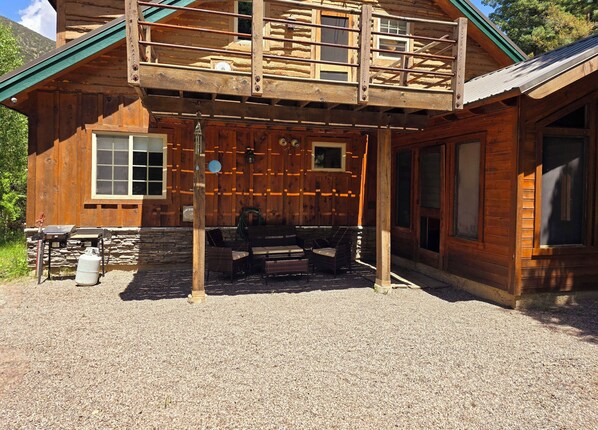 Una cabaña de madera con un porche cubierto, cimientos de piedra y un espacio para comer con mobiliario exterior.