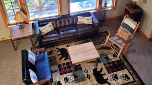 A cozy room with a leather sofa, a rocking chair, and a rug with animal prints. There are windows with a view of greenery outside.