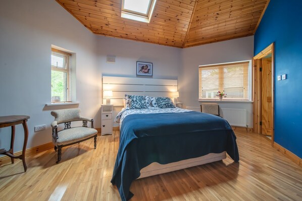 A bedroom with a wooden ceiling, a bed with a blue bedspread, a chair, a bedside table, and a picture on the wall. - Spacious 2 bed stone cottage in the Wicklow Mountains (DONARD)
