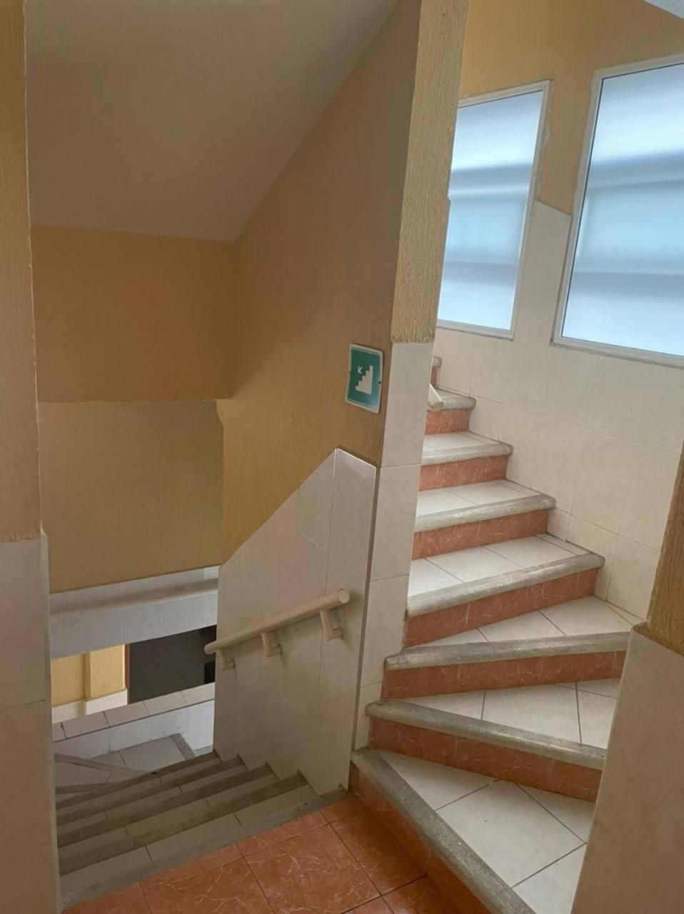 Uma escadaria com corrimão, um sinal de saída de emergência fixado na parede e uma janela com persianas.