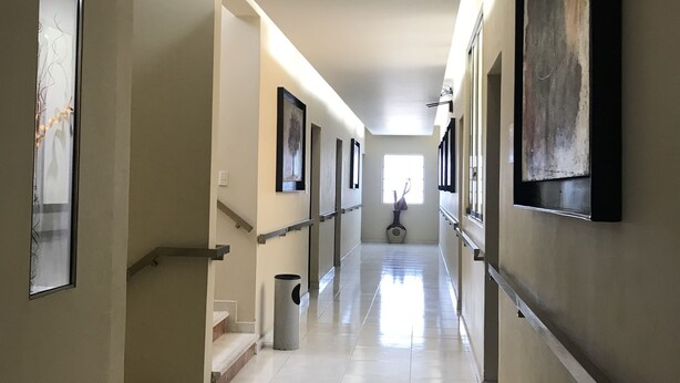 A long, well-lit hallway with tiled floors, a staircase on the left, and framed artwork on the walls.