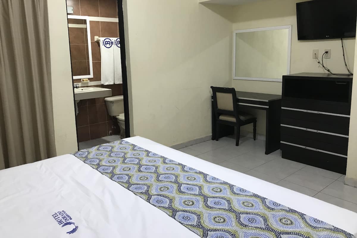 A hotel room with a bed, a desk, a chair, a dresser, a television, and a bathroom visible through an open door.