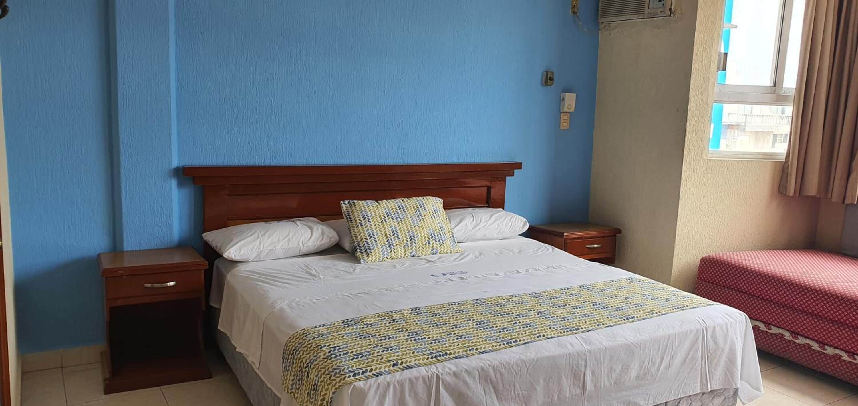 A bedroom with a wooden headboard, a bed with white bedding and a patterned throw, a bedside table, and a red bench.
