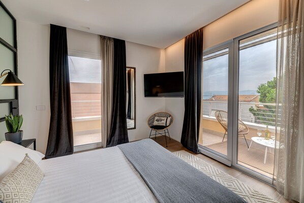 A modern hotel room with a large bed, a flat-screen TV mounted on the wall, a small seating area with a chair and table, and a balcony with a view of the city.