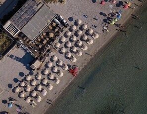 An aerial view of a beachside hotel with rows of beach umbrellas, a pool area, and a building with a sloped roof.