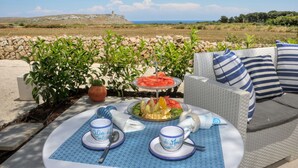 Una mesa de comedor con una bandeja de frutas, dos tazas y un plato con una cuchara, dispuesta al aire libre con un muro de piedra y plantas al fondo.
