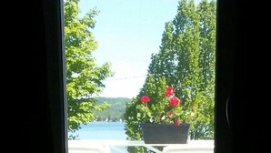 Blick von einem Zimmer auf einen Balkon mit weißer Brüstung, einen Blumentopf mit roten Blüten und ein Gewässer im Hintergrund.