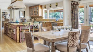 A spacious kitchen with a large island, wooden cabinets, and a dining area with a marble table and cushioned chairs. - Luxury Villa: Pool, Hot Tub, Pickleball, Ping-Pong & Bocce. 10 min to downtown! (Paso Robles)