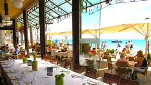 Area pranzo all'aperto con tavoli apparecchiati per gli ospiti, con vista su una spiaggia e una barca all'orizzonte.