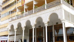 Un edificio con archi e colonne imbiancati e decorati, un balcone con ringhiera a reticello e posti a sedere all'aperto per gli ospiti.