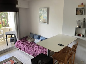 Une pièce comprenant un lit, une table, une chaise, une petite étagère avec des objets décoratifs et un tableau encadré au mur.